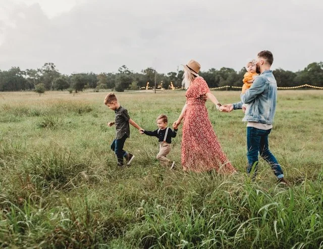 Two adults and three children walk side by side across a wide field, holding hands and staying close, creating a sense of safety and steady support.