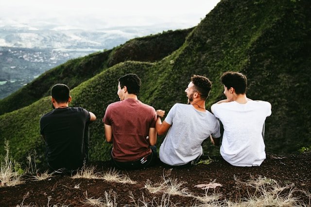 Four people sitting close together on a grassy hill, looking toward a wide, green mountain landscape, creating a sense of calm and companionship.