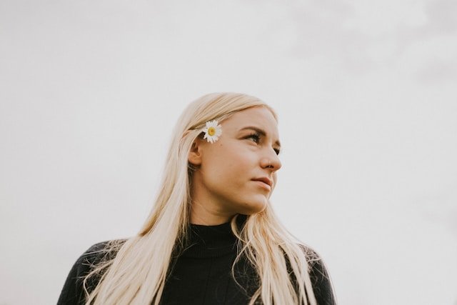 Close-up of a person with a daisy behind their ear, set against a soft, overcast sky, conveying calm and simplicity.