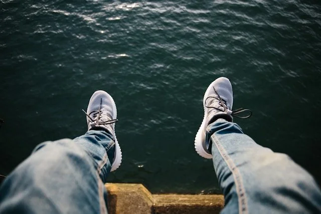 Point‑of‑view shot of legs in jeans and sneakers dangling above quiet, rippling water, creating a sense of stillness.