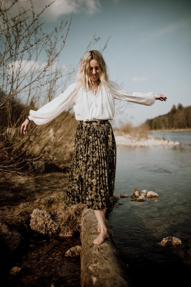 A person balances barefoot on a fallen tree over a shallow stream, arms outstretched for stability.
