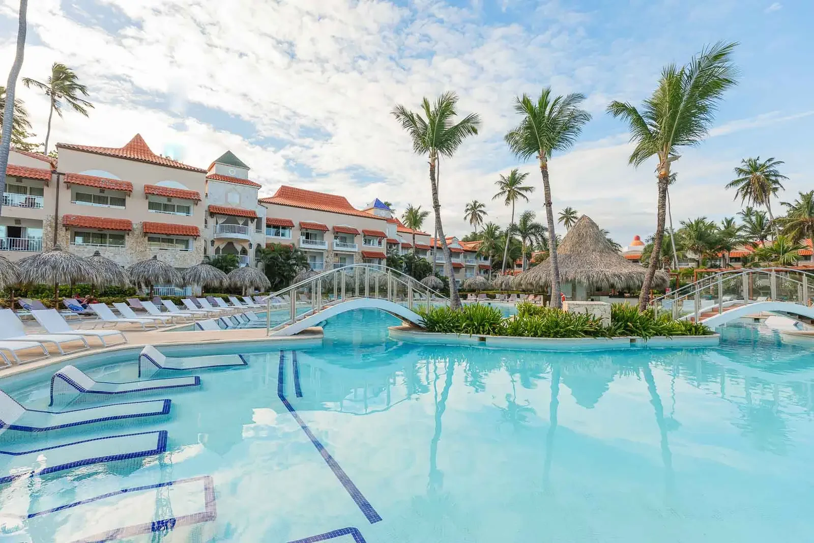 Resort swimming pool with lounge chairs, shaded huts, palm trees, and a building with red rooftops in the background under a partly cloudy sky.