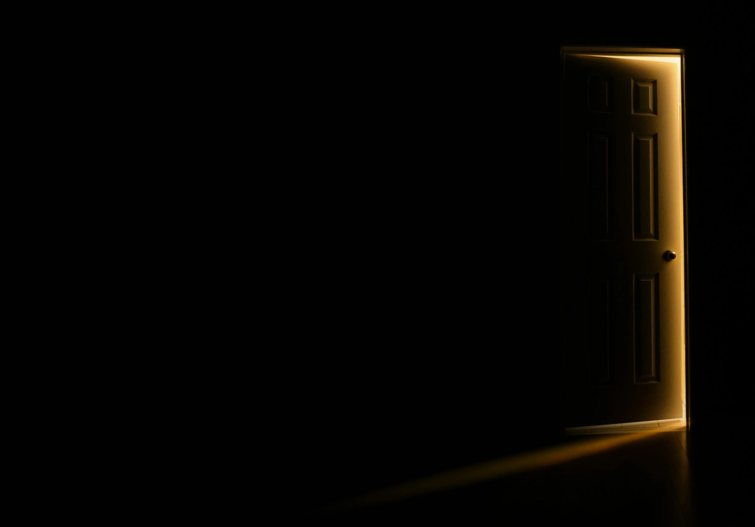 A partially open door with light shining through the gap, illuminating the floor in a dark room.