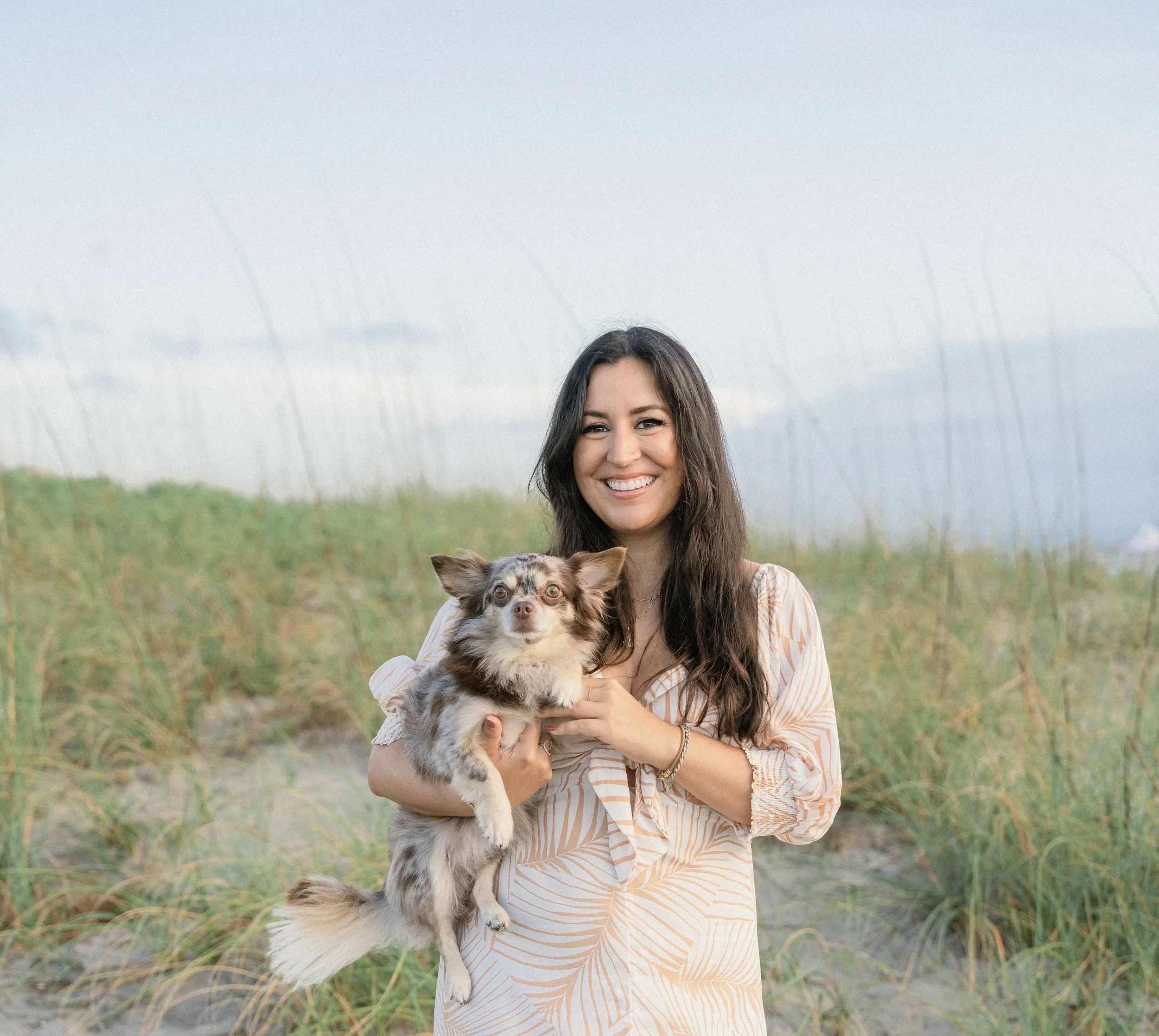 A woman with long dark hair smiling while holding a small, fluffy, merle-colored dog with upright ears, standing on a sandy, grassy beach with a blue sky and distant horizon in the background.