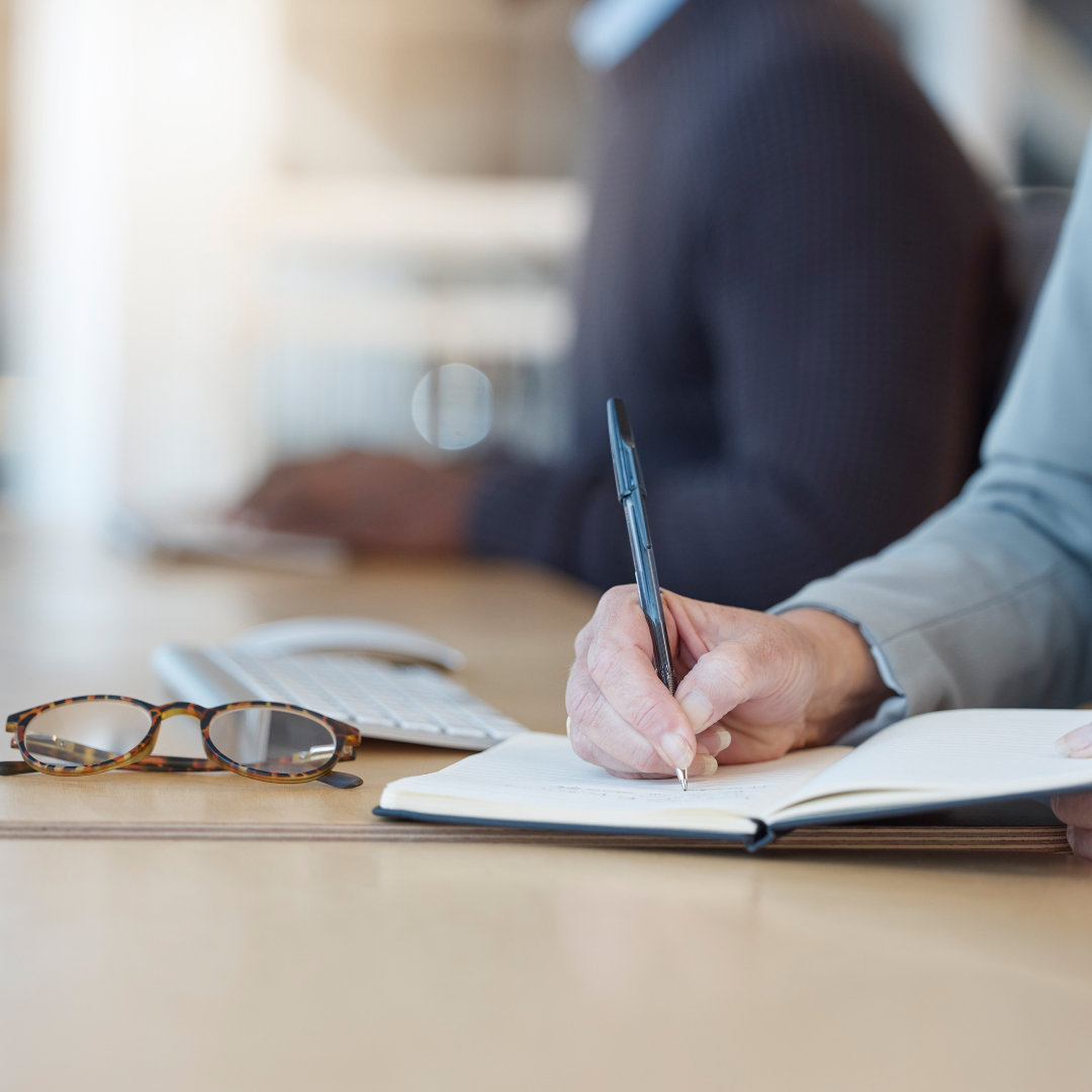 Person writing in a notebook with glasses, keyboard, and mouse on desk