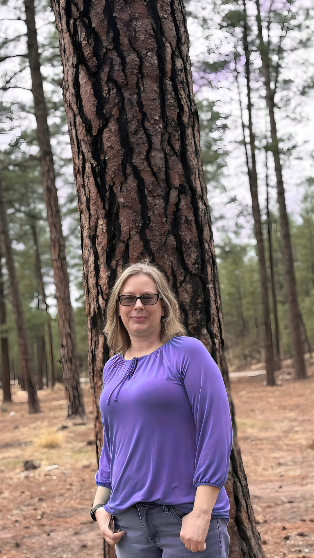 A woman with blonde hair, glasses, and a purple blouse standing in front of a large tree in a forest.