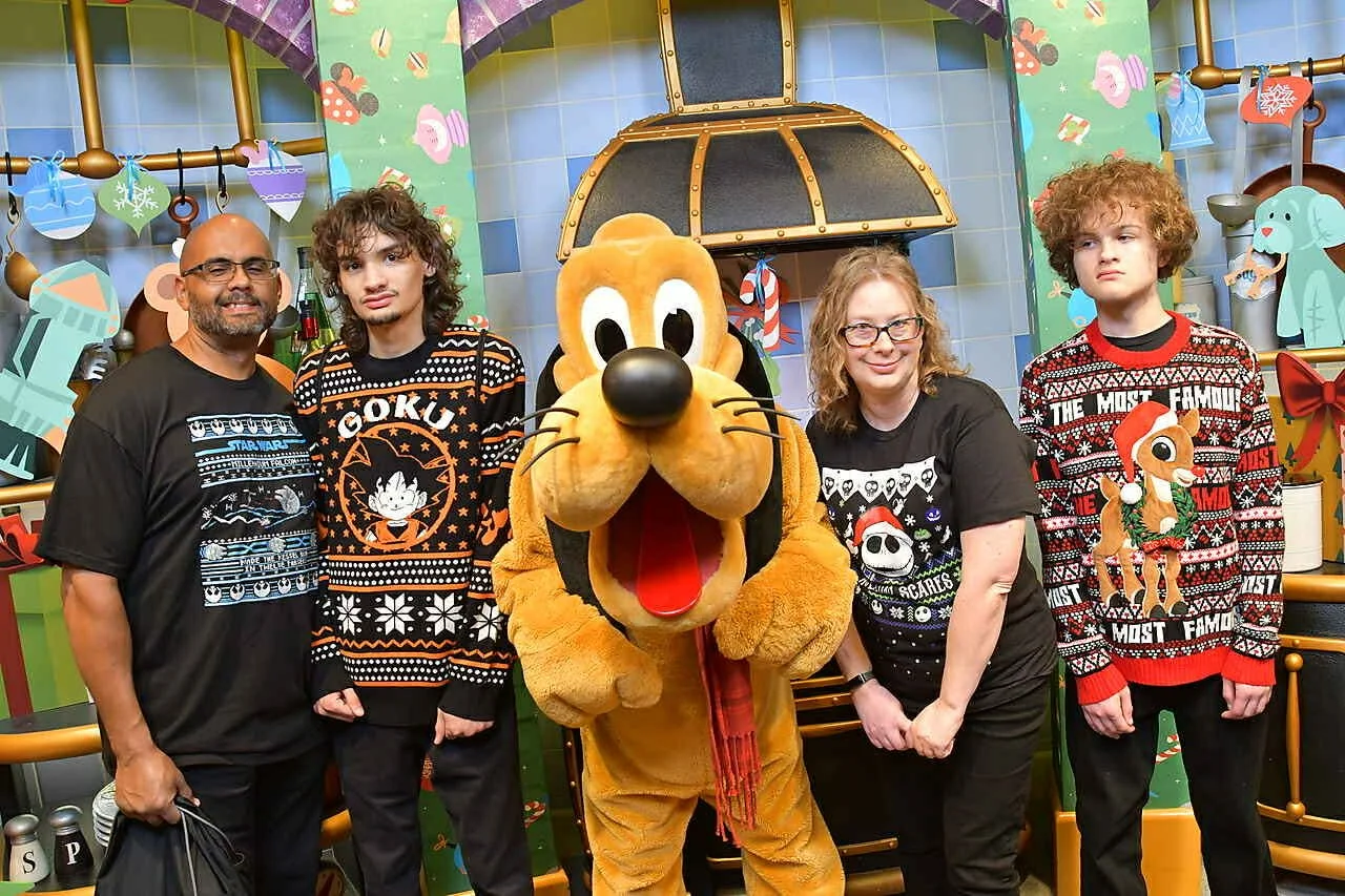 Four people and a character dressed as Pluto the dog standing in a festive, decorated setting. The people are wearing Christmas-themed sweaters, and the background features holiday decorations and toys.