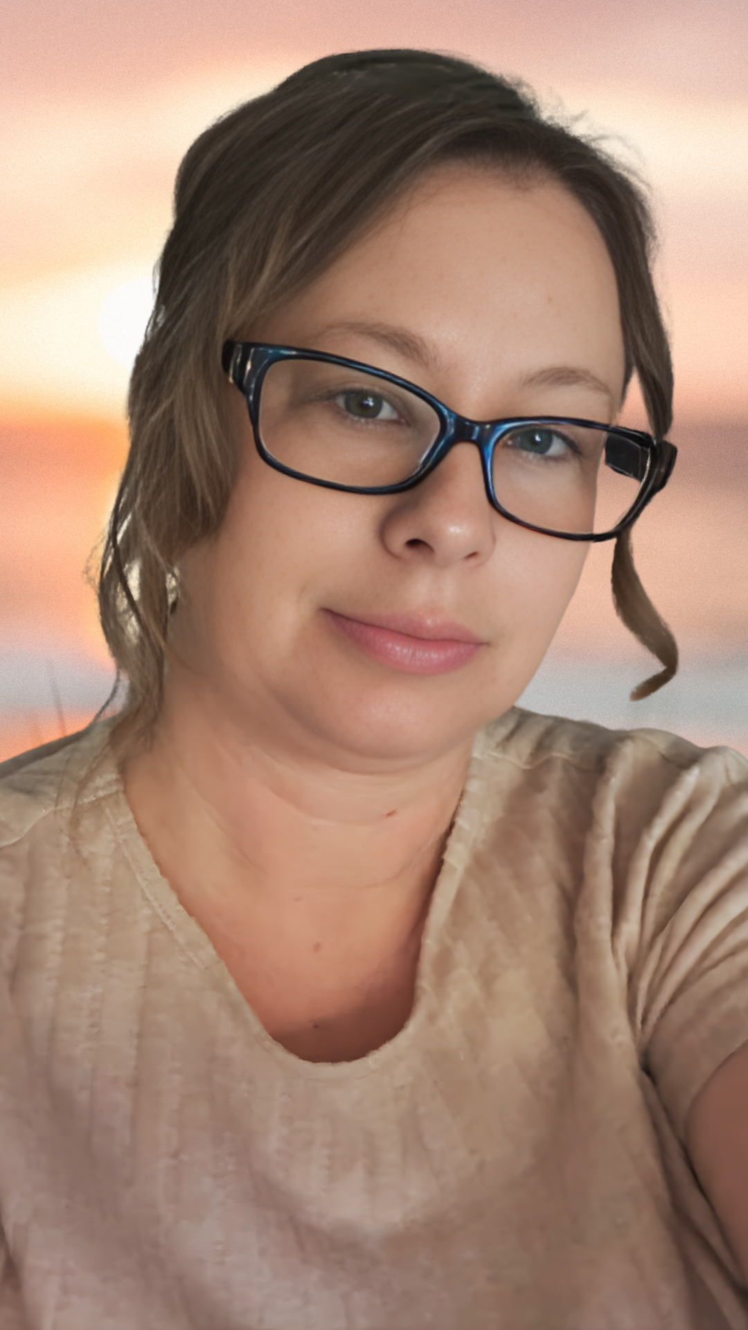 A woman with short brown hair and glasses taking a selfie with a sunset blurred background.