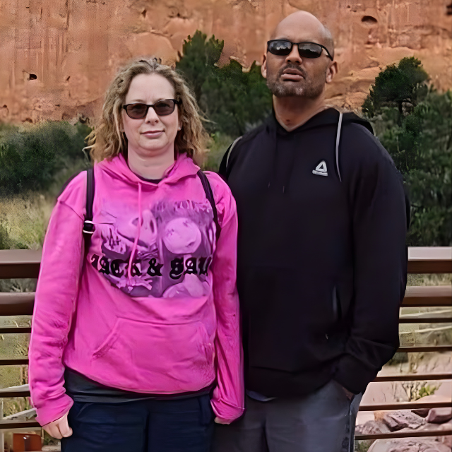 A woman and a man standing outdoors near a wooden railing with a red rock cliff and green trees in the background, both wearing sunglasses.