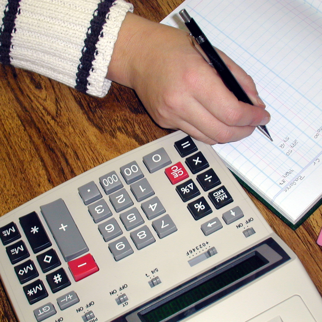 A person's hand writing in a grid notebook next to a calculator on a wooden desk.