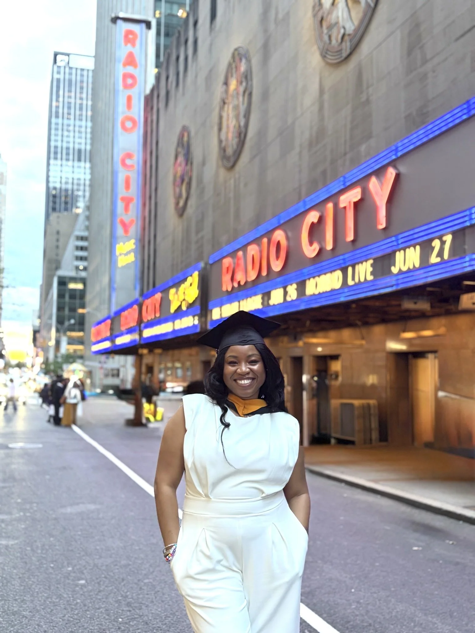 Smiling woman in white outfit standing on city street with Radio City marquee in background.