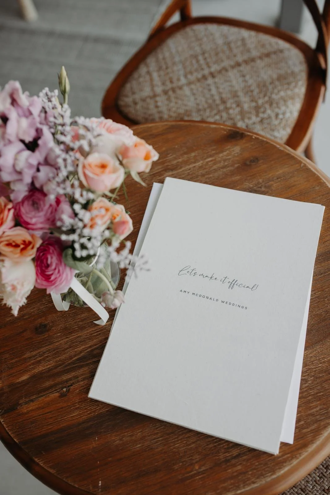 A wedding vow book titled 'Let's make it official!' placed on a round wooden table, next to a colorful bouquet of pink, peach, and white flowers with greenery.