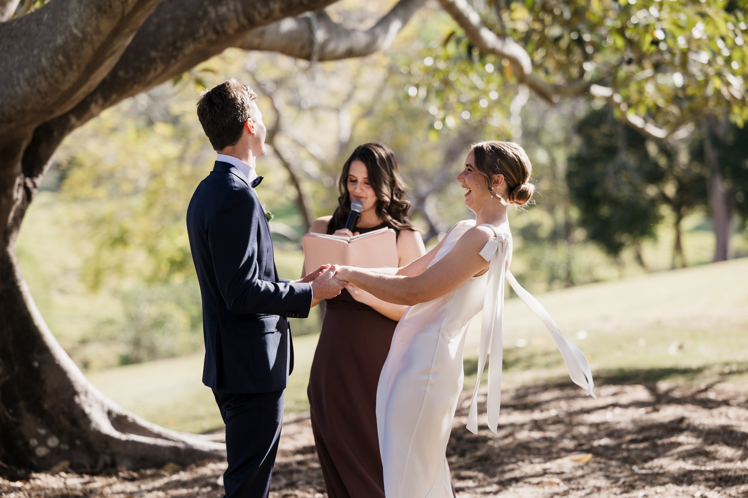 A couple getting married outdoors, holding hands and smiling at each other, with Sunshine Coast Civil Celebrant Amy McDonald standing behind them delivering the ceremony, surrounded by trees and sunlight.