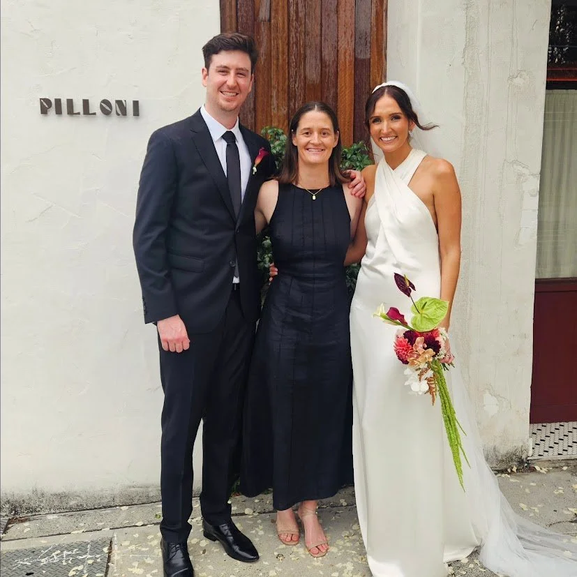 A wedding photo of three people, a man in a black suit, Sunshine Coast Civil Celebrant Amy McDonald in a black dress, and a woman in a white wedding gown holding a bouquet, standing outside Pilloni Restaurant in West End Brisbane.