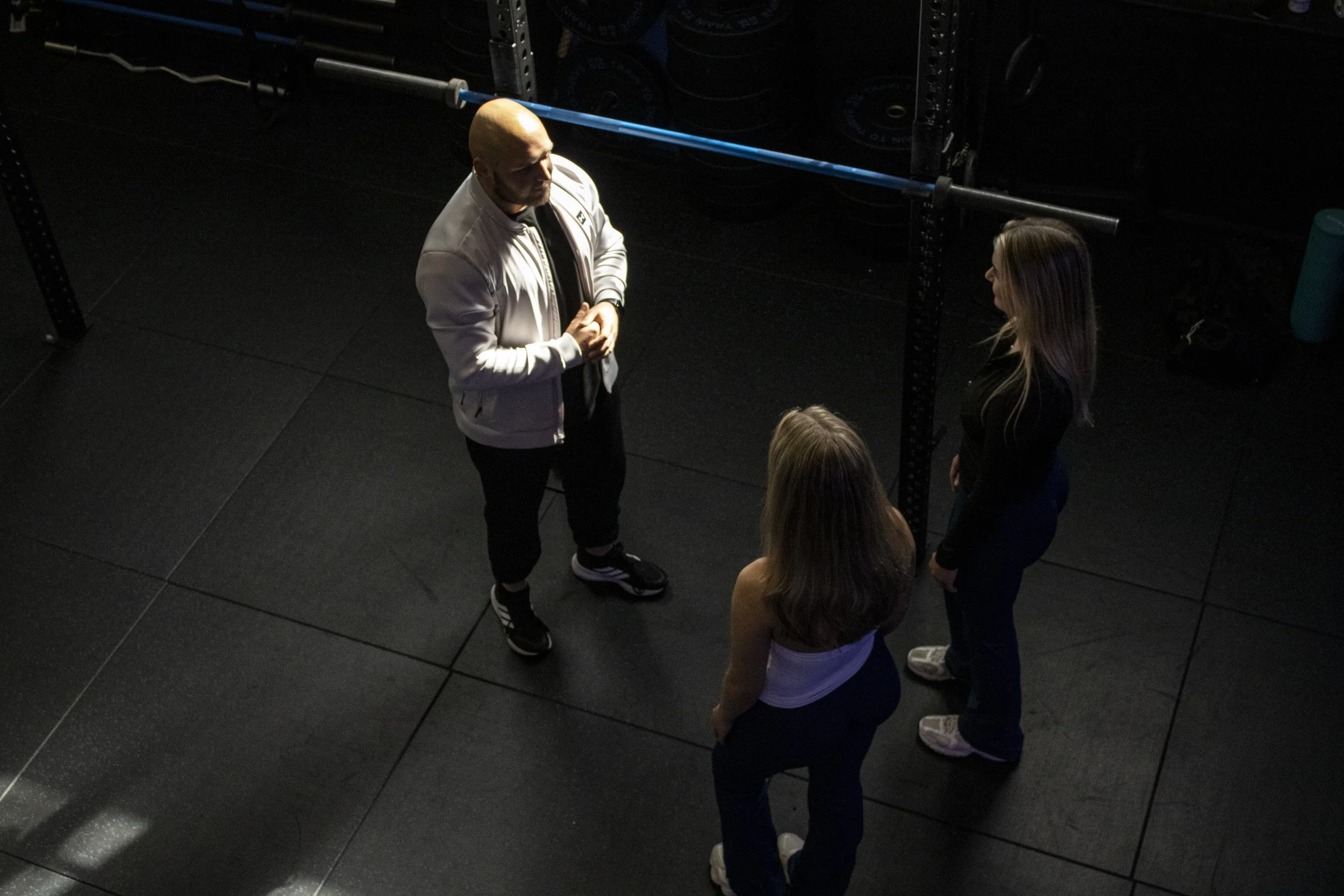 A coach and two of his clients standing by a squat rack, the coach is explaining the exercise & form of set exercise at Carlisle's Cumbria Gym in Kingstown. He is coaching two ladies.
