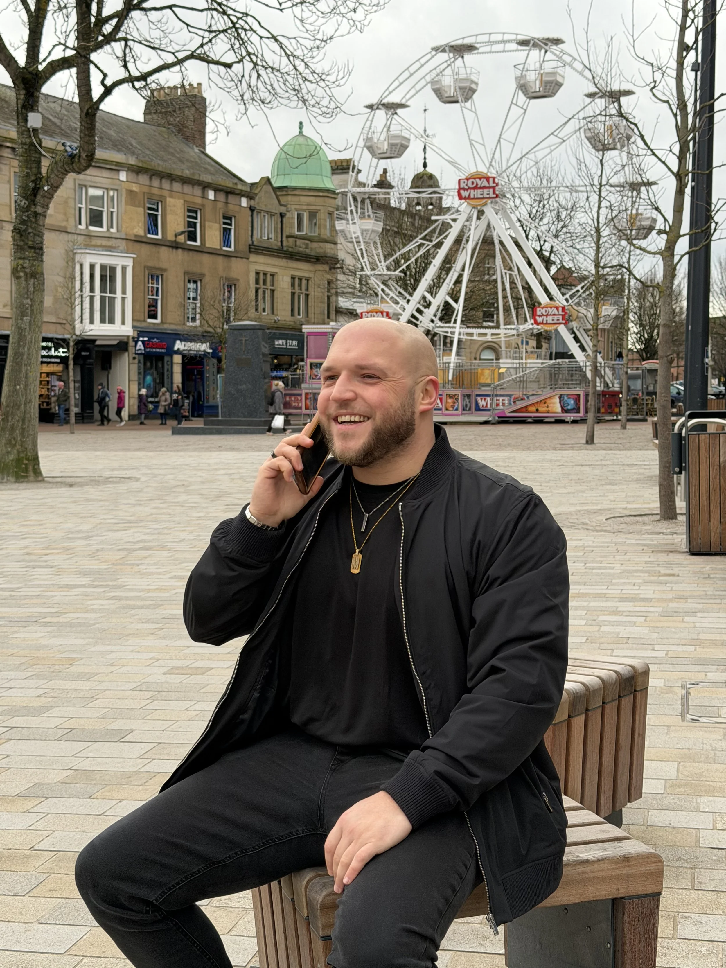 A smiling man with a bald head and beard sitting on a bench in a city square, talking on his cellphone, with a large Ferris wheel in the background.