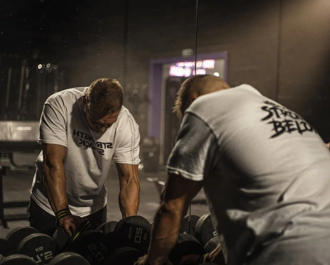 Two men lifting dumbbells in a gym, reflected in a mirror.