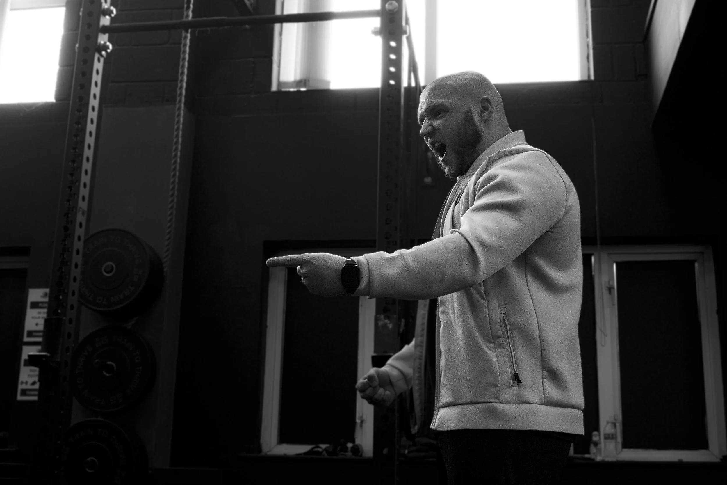 A man with a shaved head and beard, wearing a light-colored jacket, is giving an intense speech or shouting while pointing his finger inside a gym with weight racks and a black brick wall.