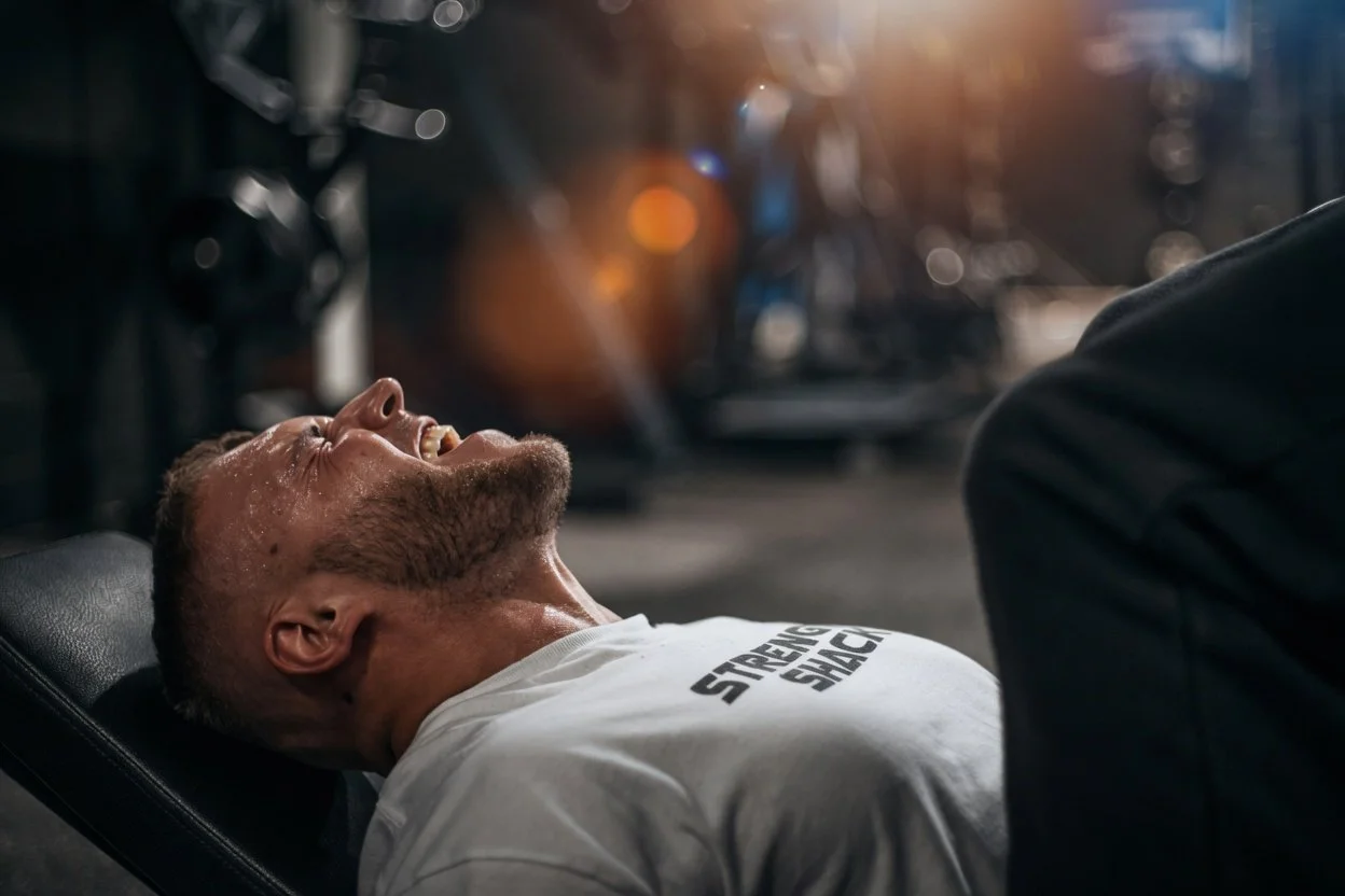 A man sweating and laughing while lying on a workout bench in a gym, wearing a white t-shirt that says 'Strength Since'.