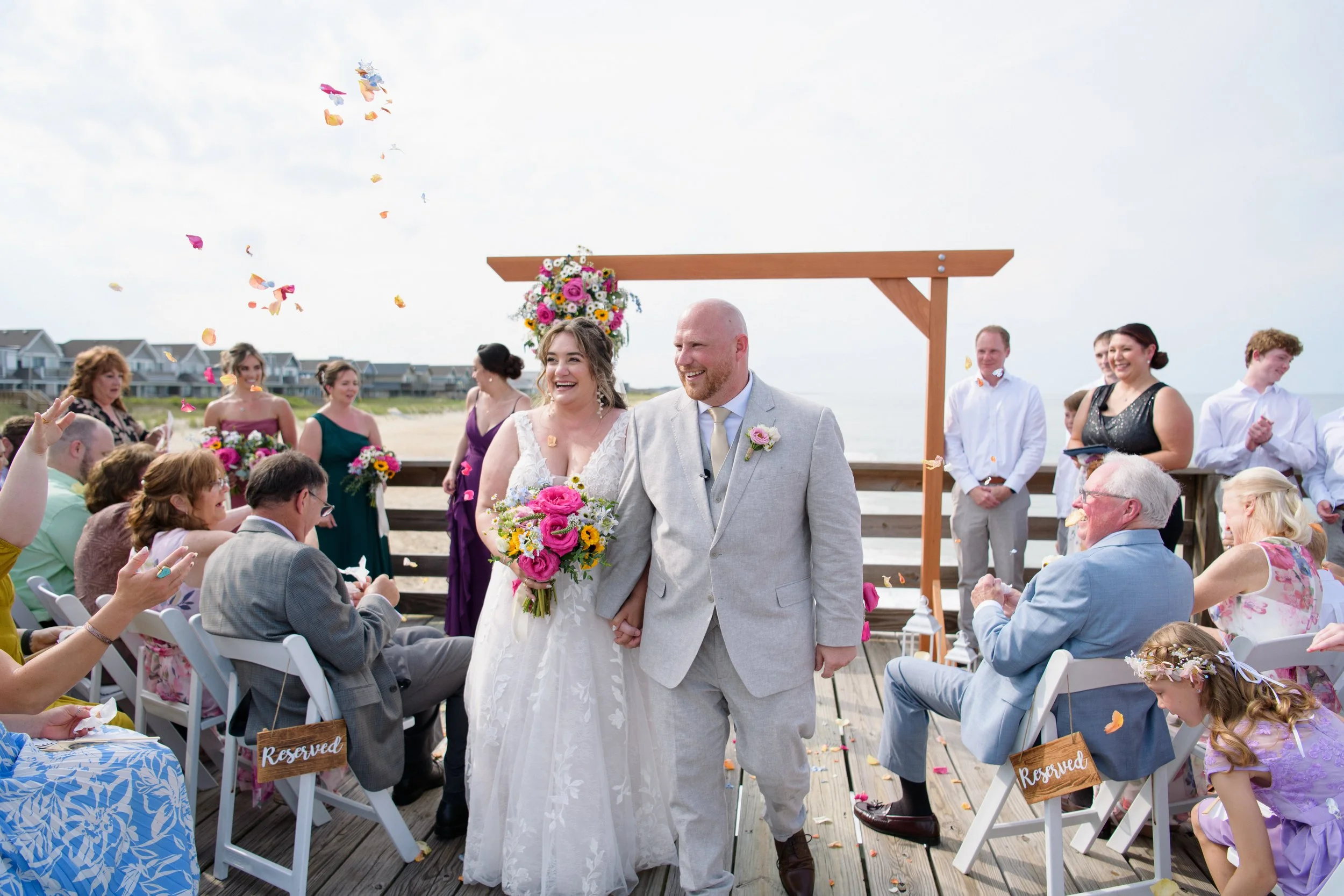 A wedding ceremony on a beach with a bride and groom walking down the aisle, surrounded by guests celebrating and throwing flower petals, with a floral arch in the background.
