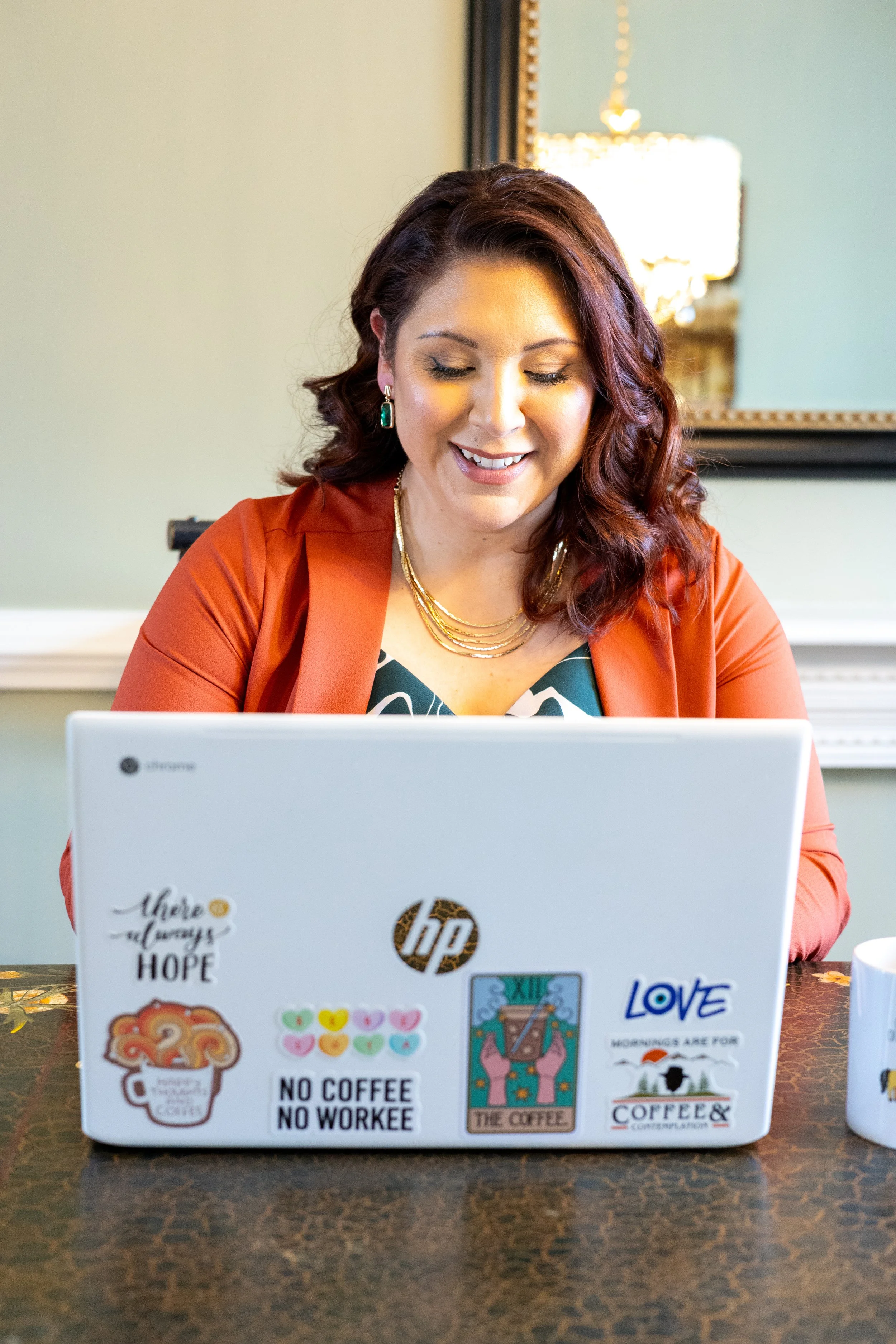 Smiling woman with wavy, reddish-brown hair using a laptop decorated with various stickers, sitting at a table in a well-lit room.