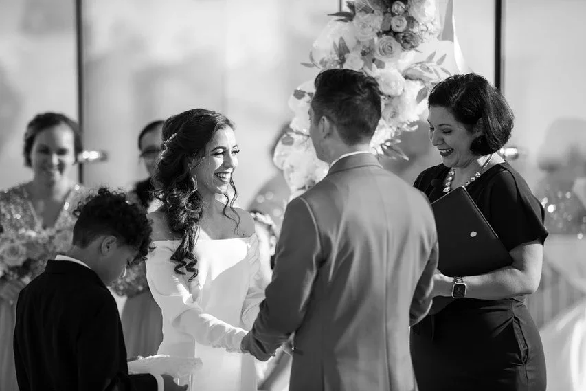 A wedding ceremony with a bride and groom holding hands, a young boy, and a woman officiating, all smiling. In the background, wedding guests and floral decorations are visible.