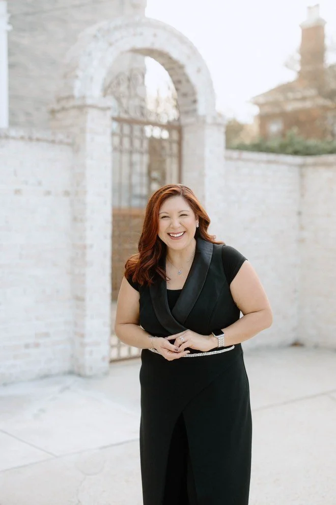 Woman with red hair in a black dress smiling outdoors in front of a brick archway.