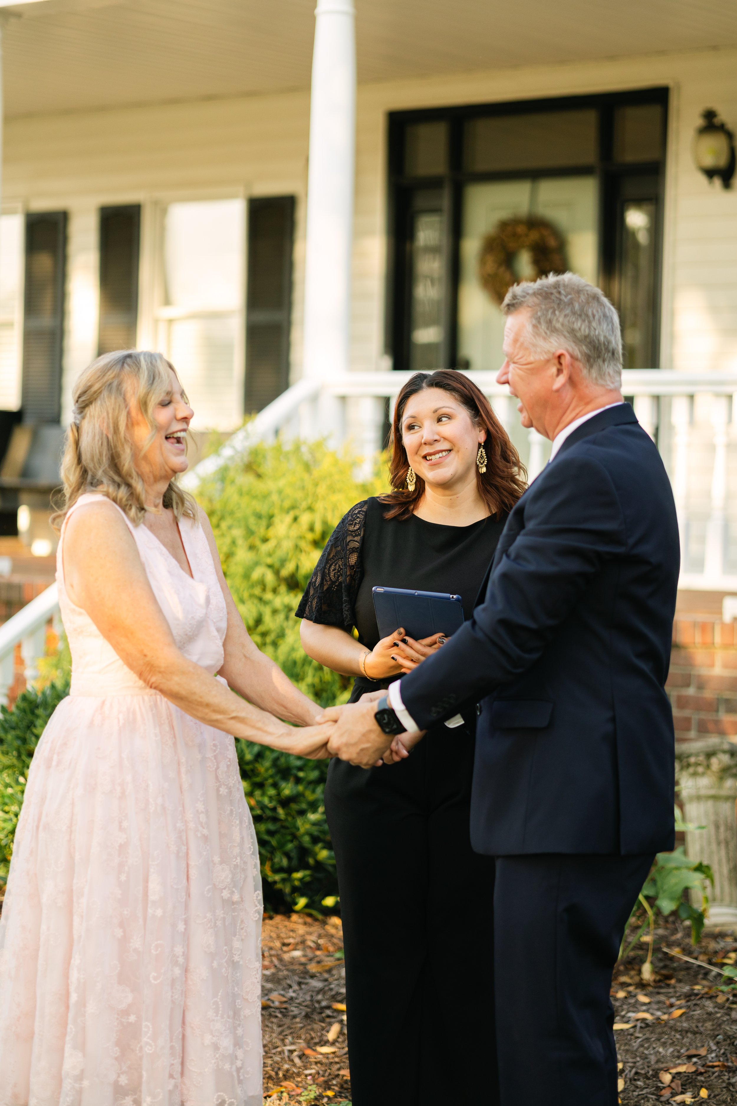 A bride and groom holding hands and smiling at each other during a wedding ceremony outside a house, with a woman officiant standing between them holding a tablet, all smiling.