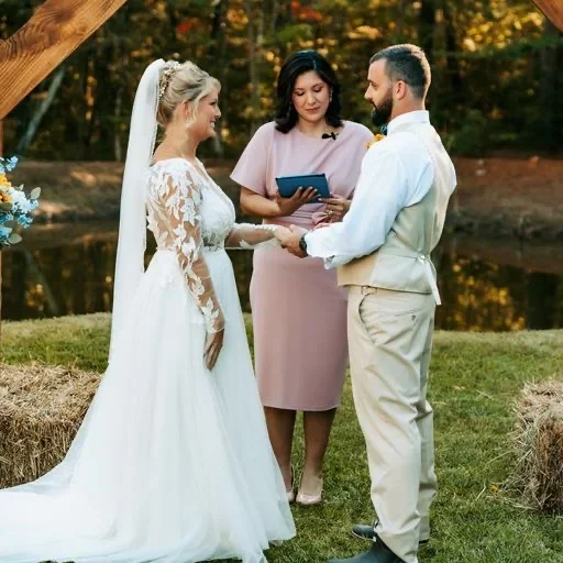 A bride and groom holding hands, standing outdoors near a pond with trees in the background, during their wedding ceremony, with an officiant holding a book between them.