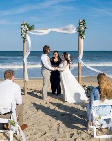 A beach wedding ceremony with a couple getting married under a wooden arch decorated with white flowers and fabric, with guests seated on wooden chairs nearby, ocean waves in the background, and a clear blue sky.