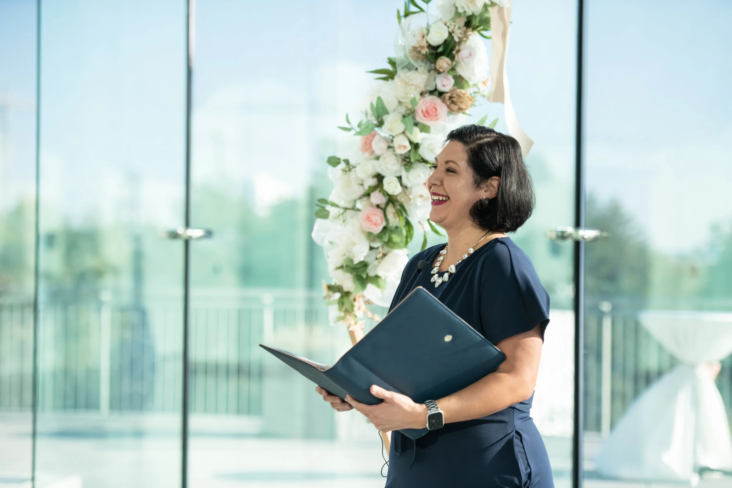 A woman in a navy blue dress holding a folder, smiling, with a floral arrangement in the background during a wedding ceremony.