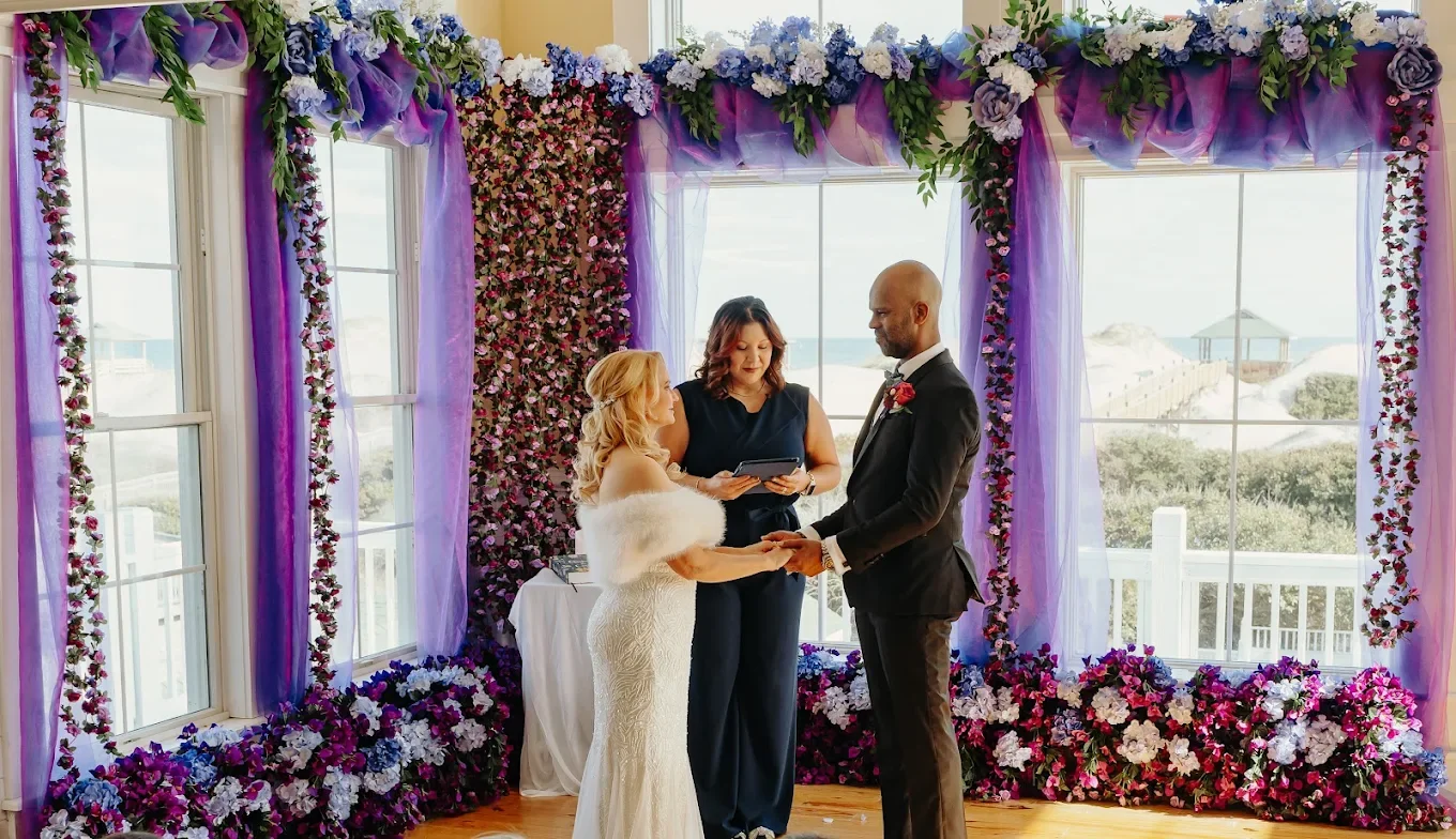 A wedding ceremony with a bride and groom holding hands and exchanging vows, standing in front of a floral backdrop with purple and pink flowers and sheer purple curtains, in a bright room with large windows showing a beach view.