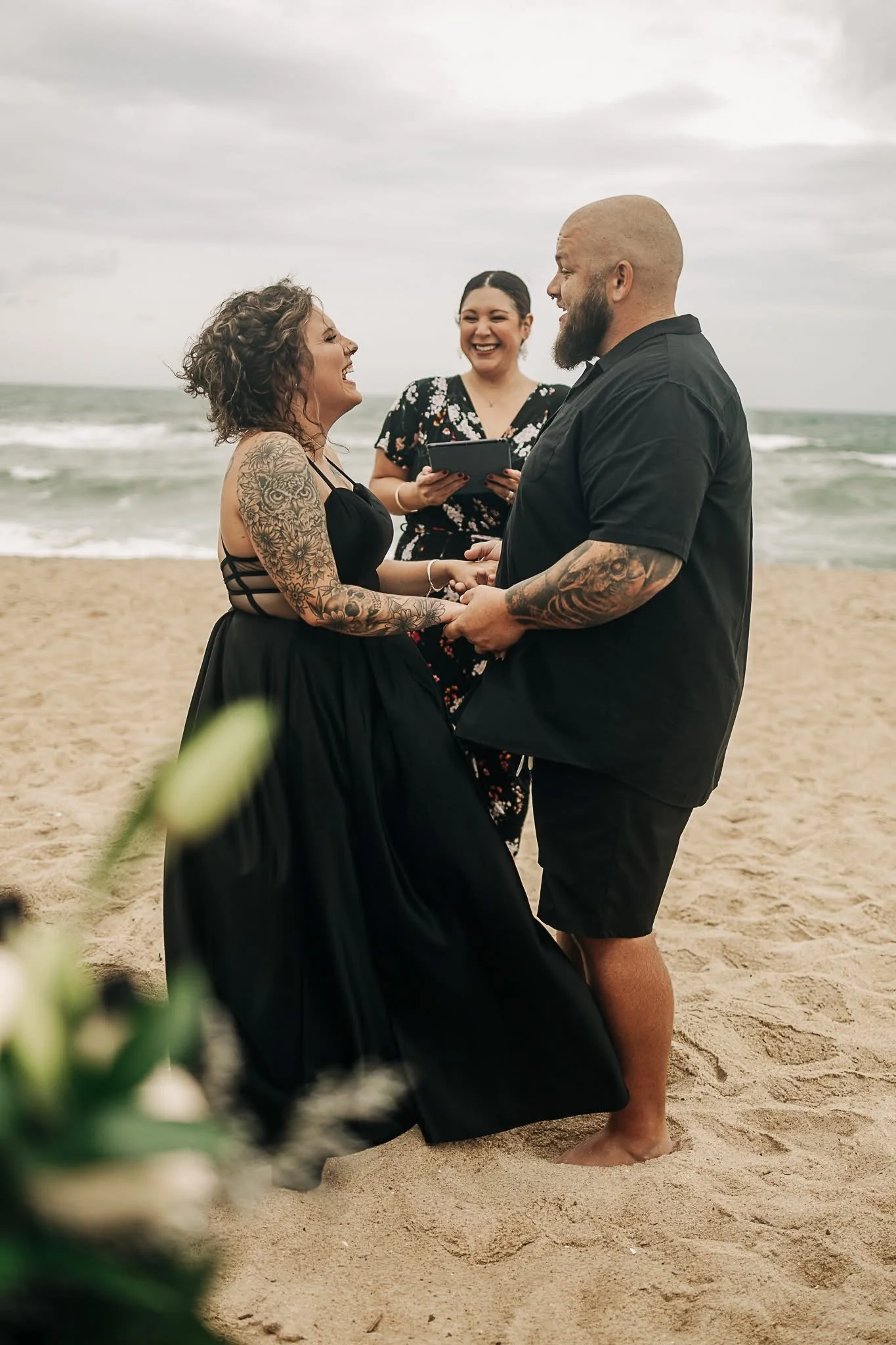 A couple gets married on a beach, holding hands and smiling at each other while a woman officiant stands behind them holding a tablet and smiling.