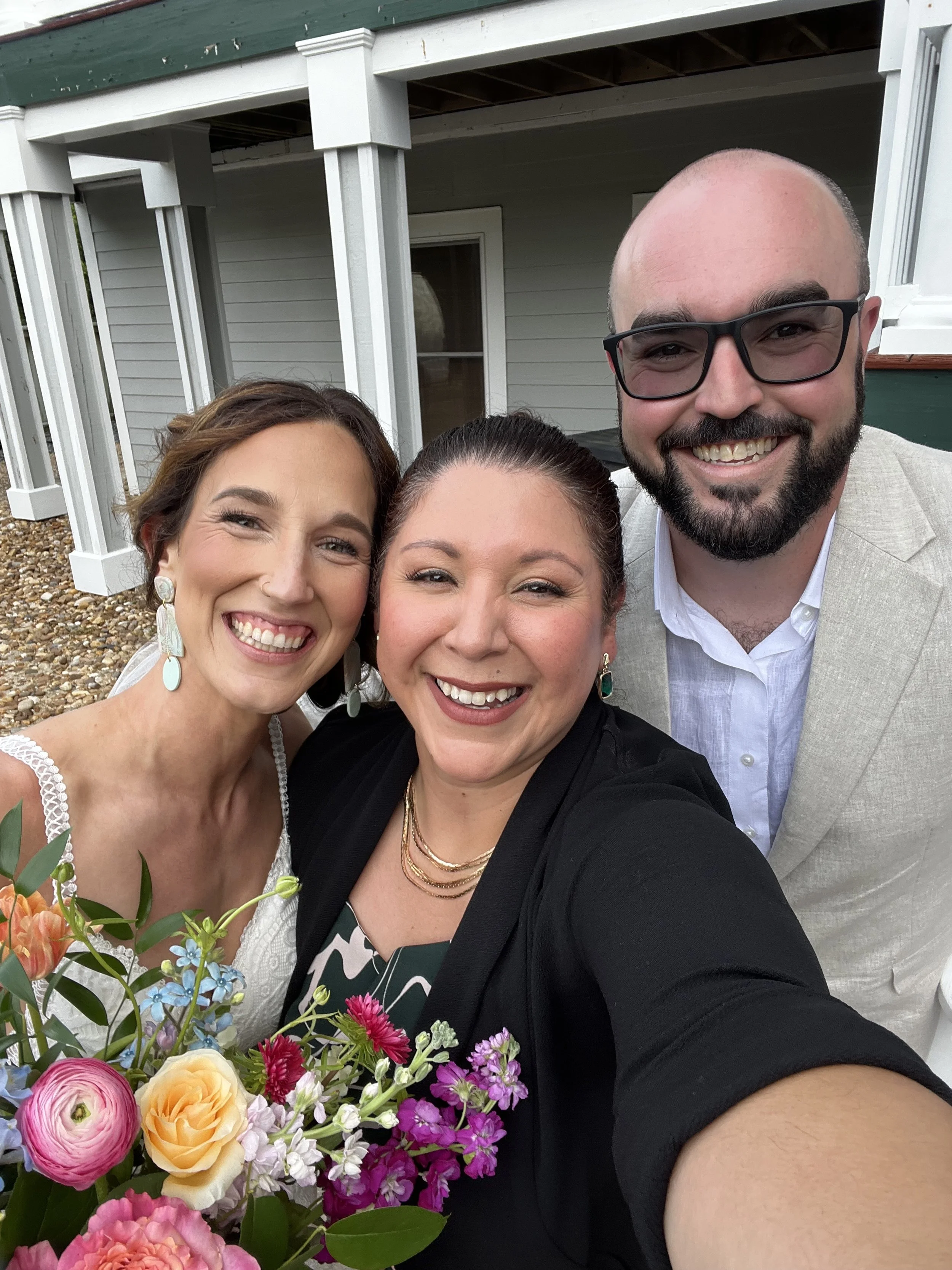Three smiling people taking a selfie outdoors near a house, with a woman holding a colorful bouquet of flowers in the foreground.