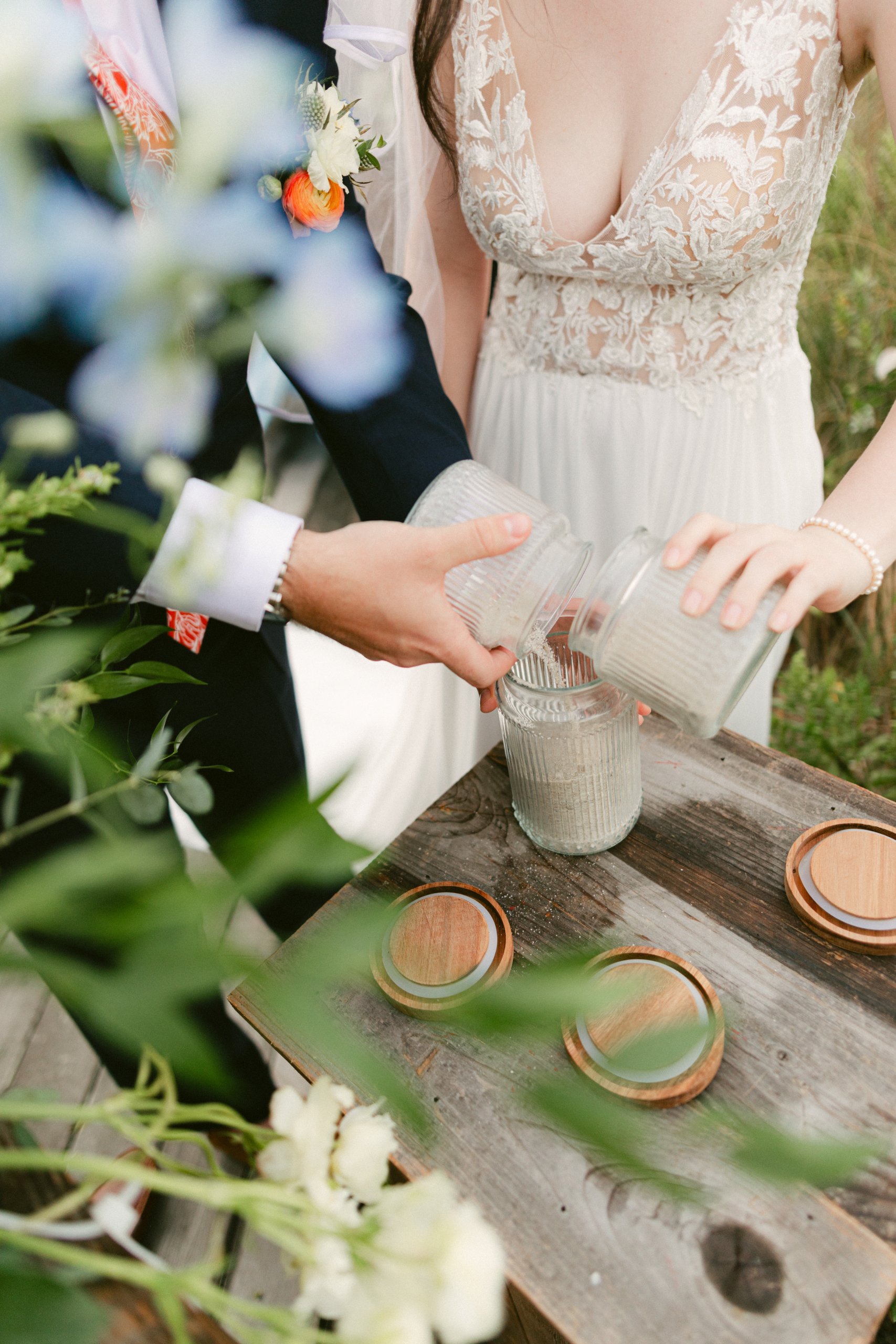 A bride and groom pour sand from glass jars into a larger container during a sand ceremony at their wedding, with greenery and wedding accessories visible.