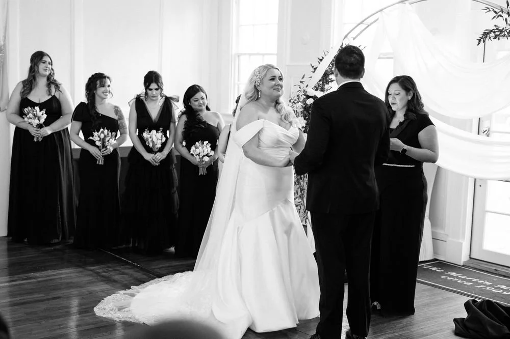 A wedding ceremony with a bride and groom exchanging vows, five bridesmaids holding bouquets, and a female officiant in an indoor setting with natural light.