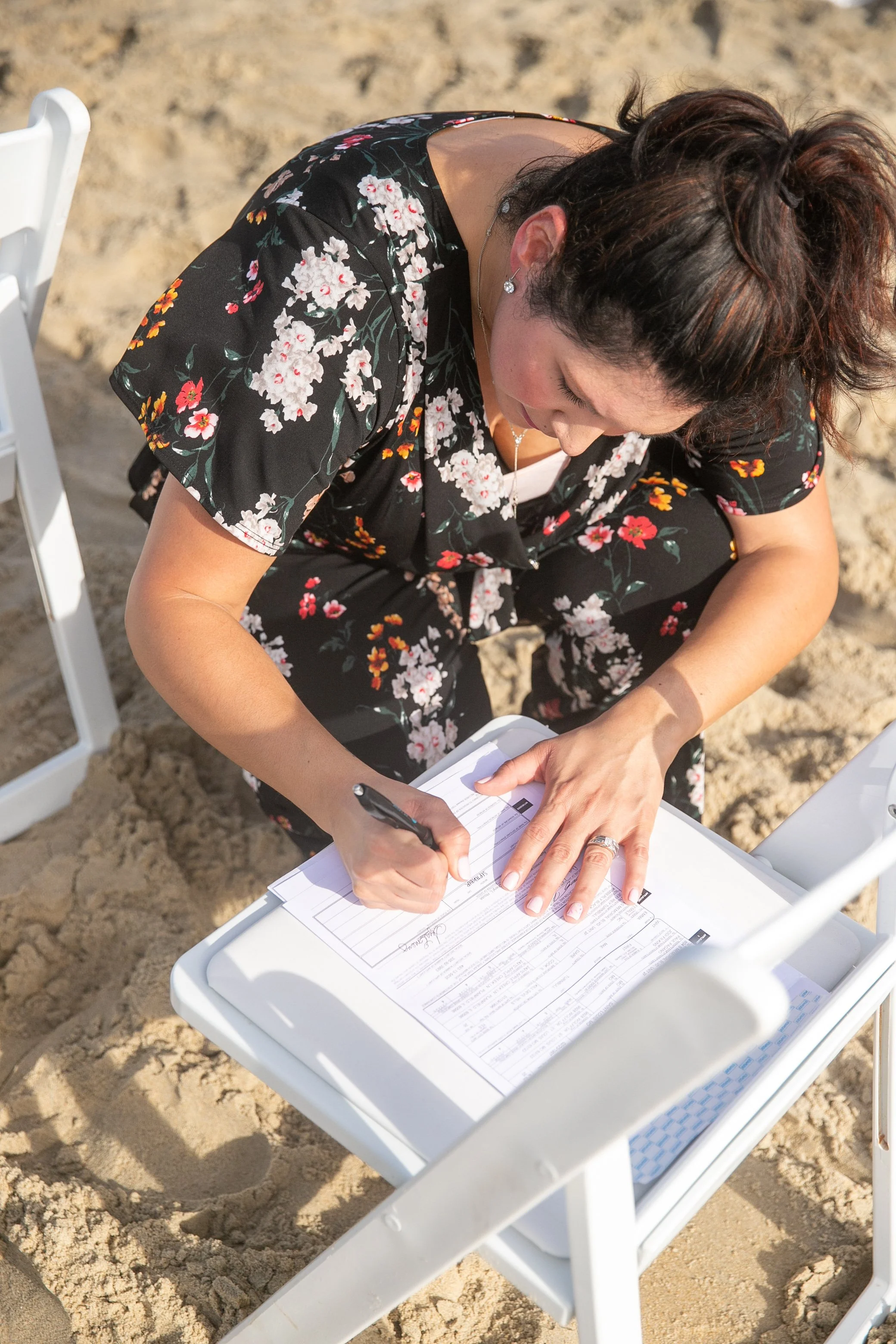 A woman in a black floral dress sitting on a white chair on a sandy beach, writing on a clipboard.