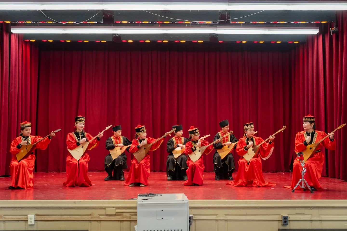 Group of children dressed in traditional costumes performing on stage with stringed instruments in front of red curtain.