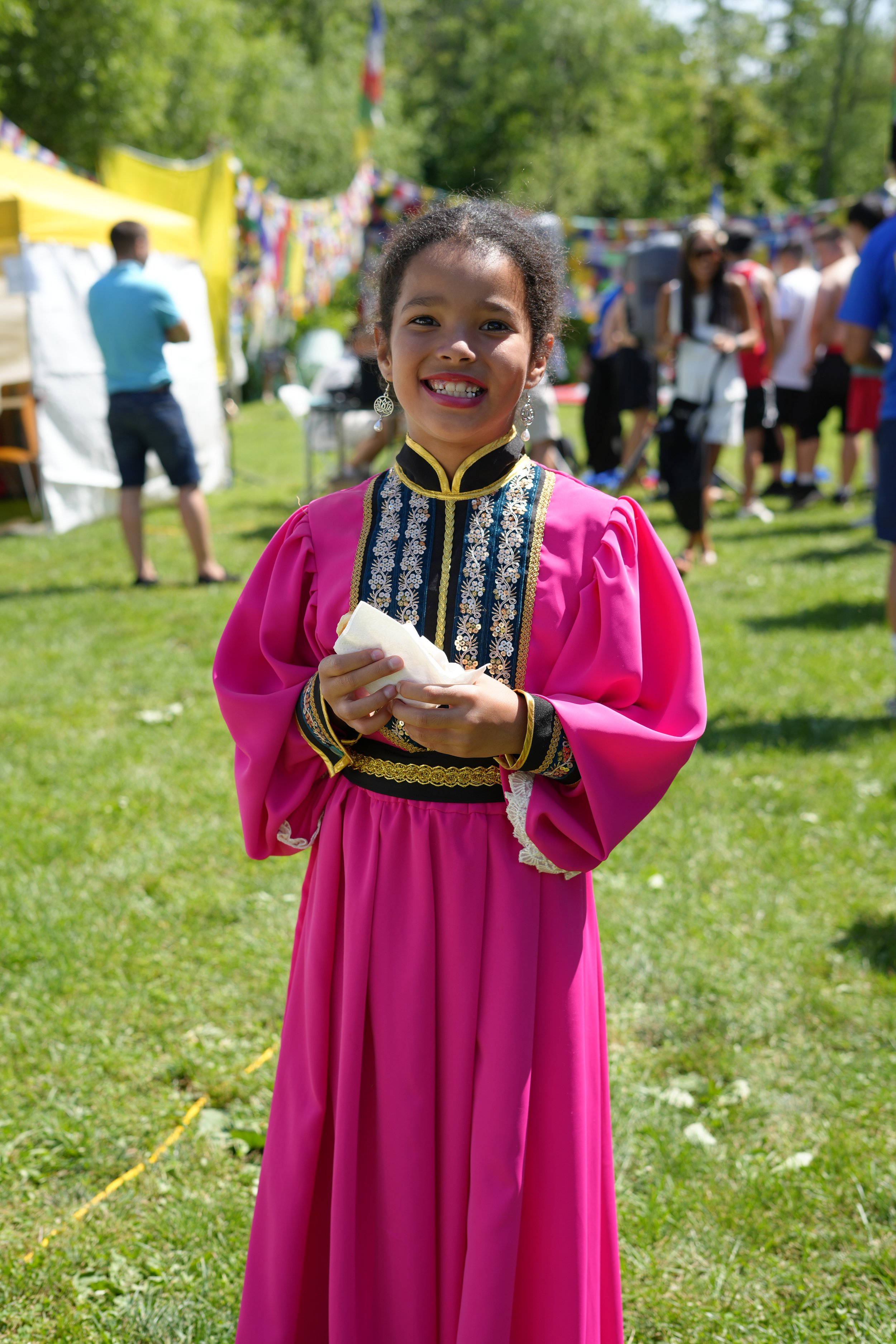 Smiling young girl in a traditional bright pink dress with black and gold accents at an outdoor festival or celebration.