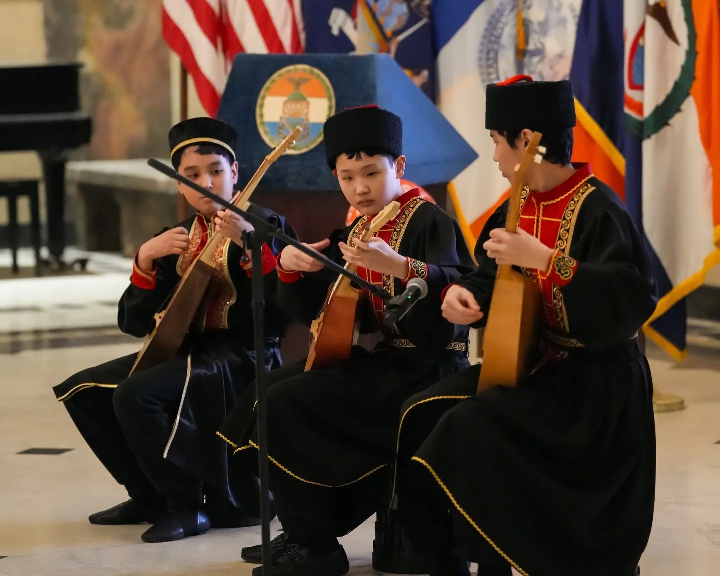 Three boys in traditional attire playing guitars during a performance with flags and a podium in the background.