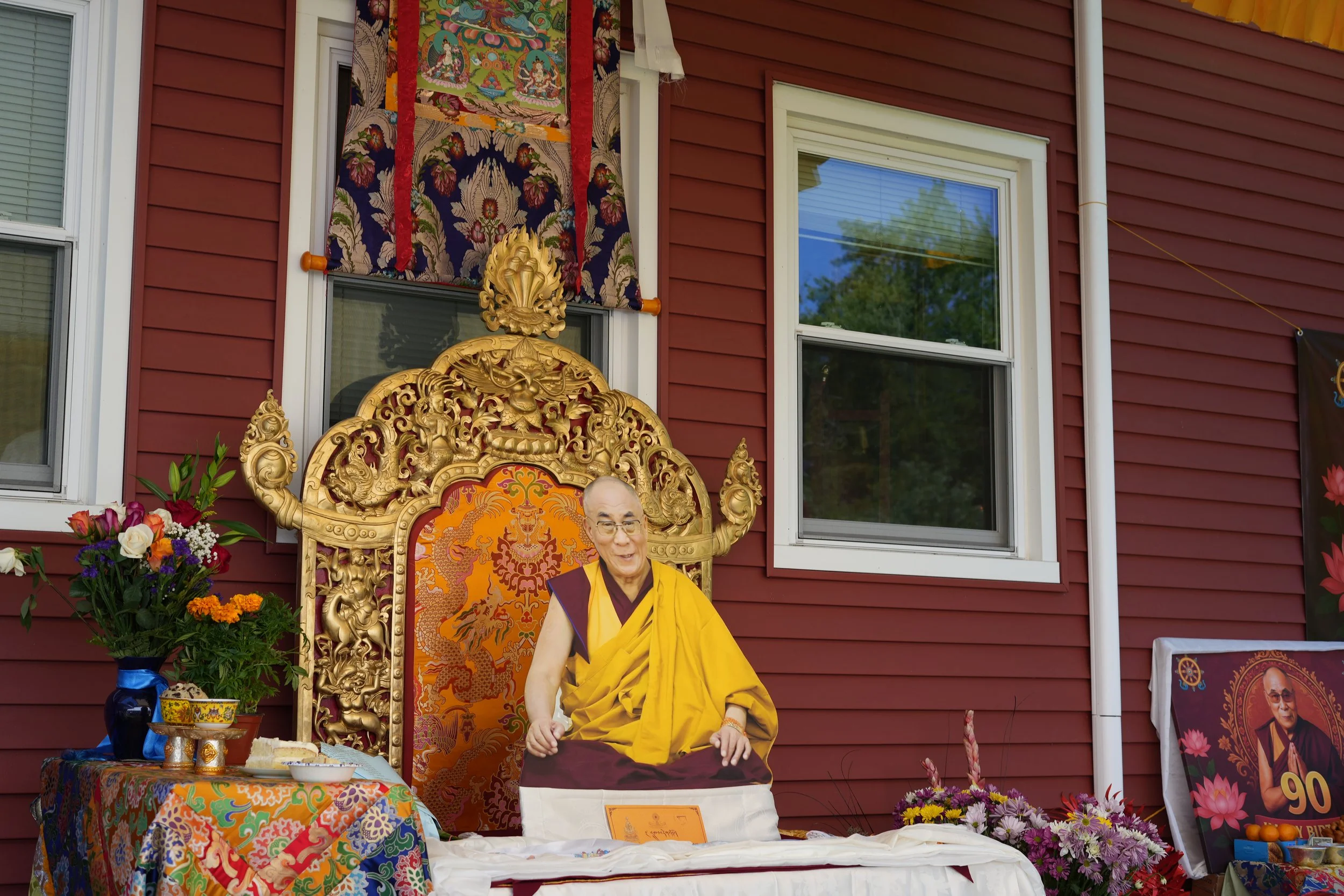 A Buddhist monk statue seated on an ornate golden throne outside a red house, with flower arrangements and a commemorative poster nearby.