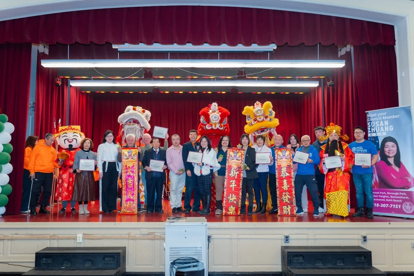 Group of people on stage in front of red curtain, celebrating during a cultural event with lion dance costumes, holding certificates, and colorful banners with Chinese characters.