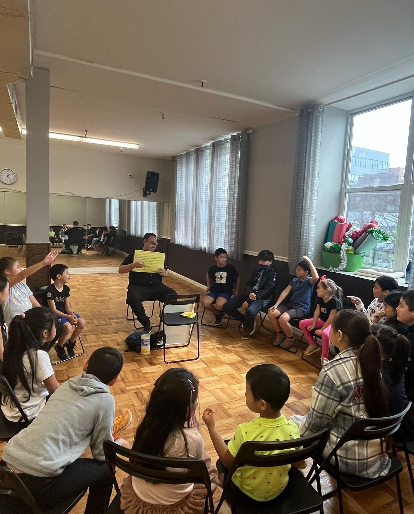 Children sitting in a circle on chairs and the floor, listening to an adult holding a yellow paper, in a room decorated with Christmas ornaments on the windowsill.