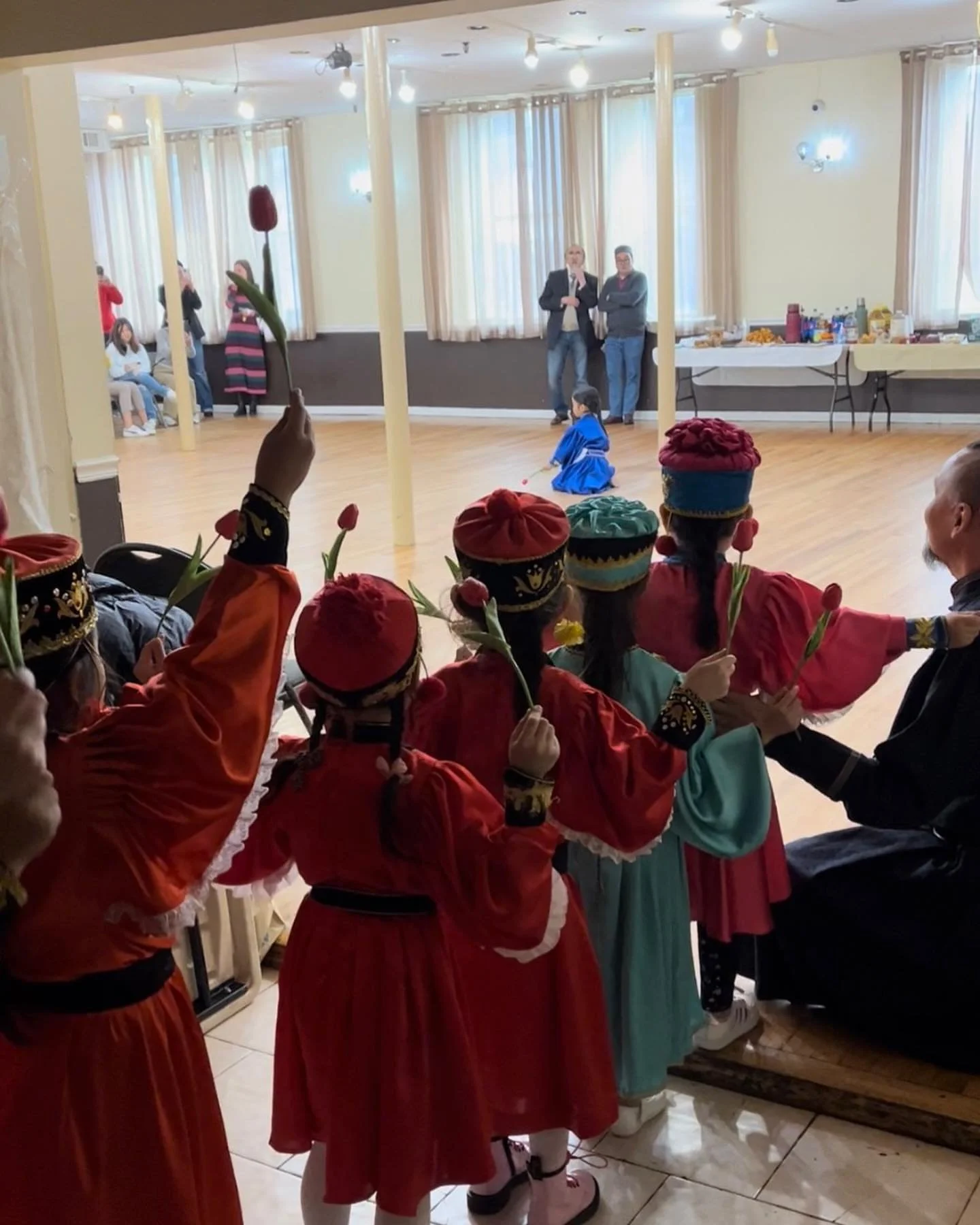 Children dressed in traditional costumes holding tulips, watching a performance at a cultural event indoors.