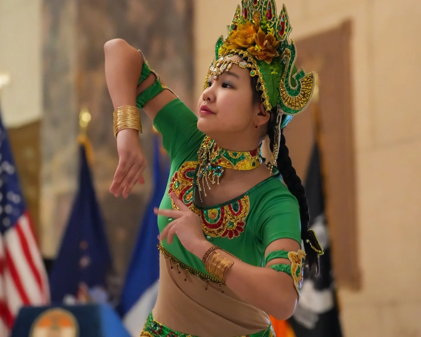 Young girl performing a traditional dance in elaborate green and gold costume with headpiece, jewelry, and braid, indoors with flags in the background.