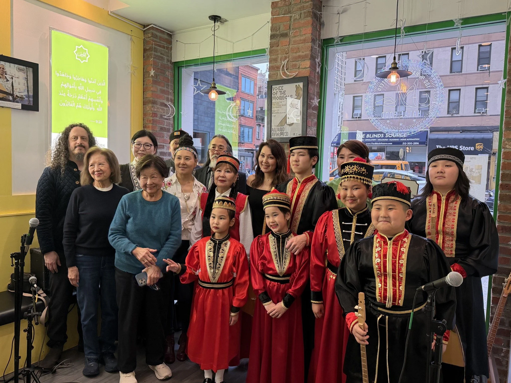 Group of people, including children in traditional costumes, gathered inside a room with brick walls and large windows, celebrating an event or cultural occasion.