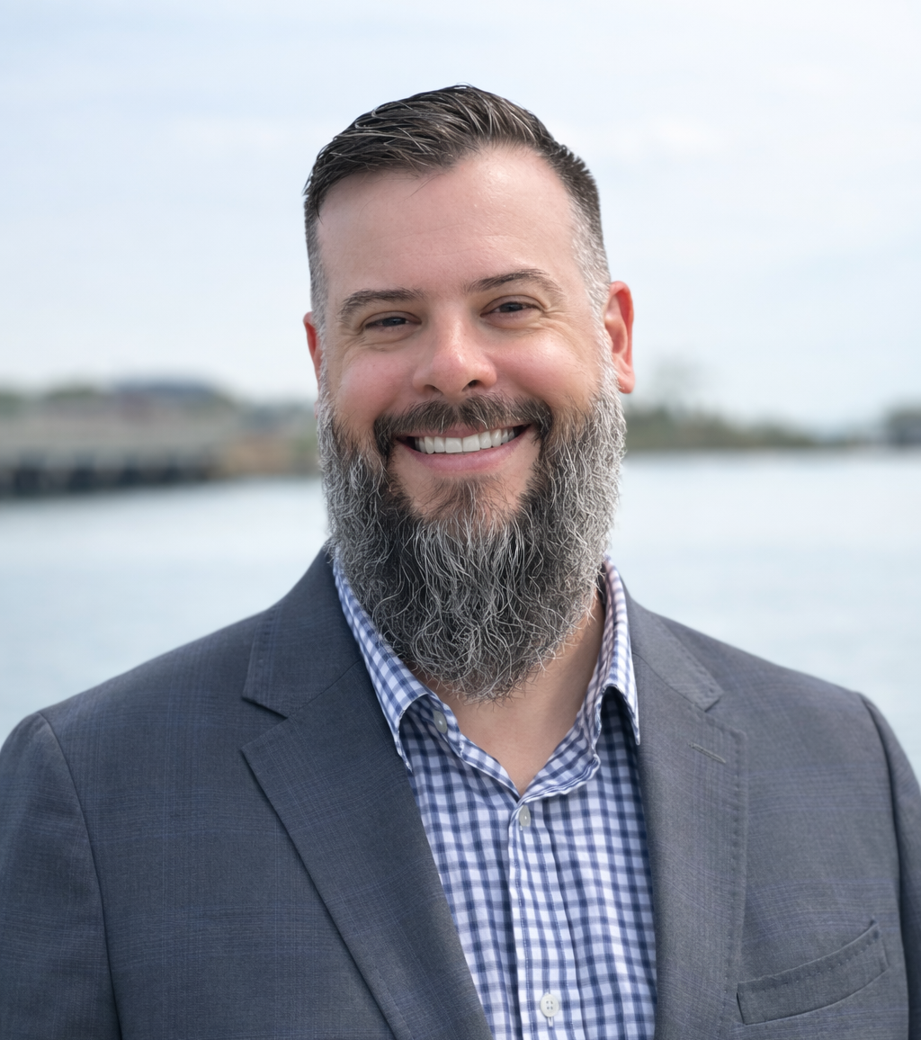 Commissioner Joe Griffith wearing a gray suit and a checkered shirt, standing outdoors in front of Mill Creek at Fort Monroe.