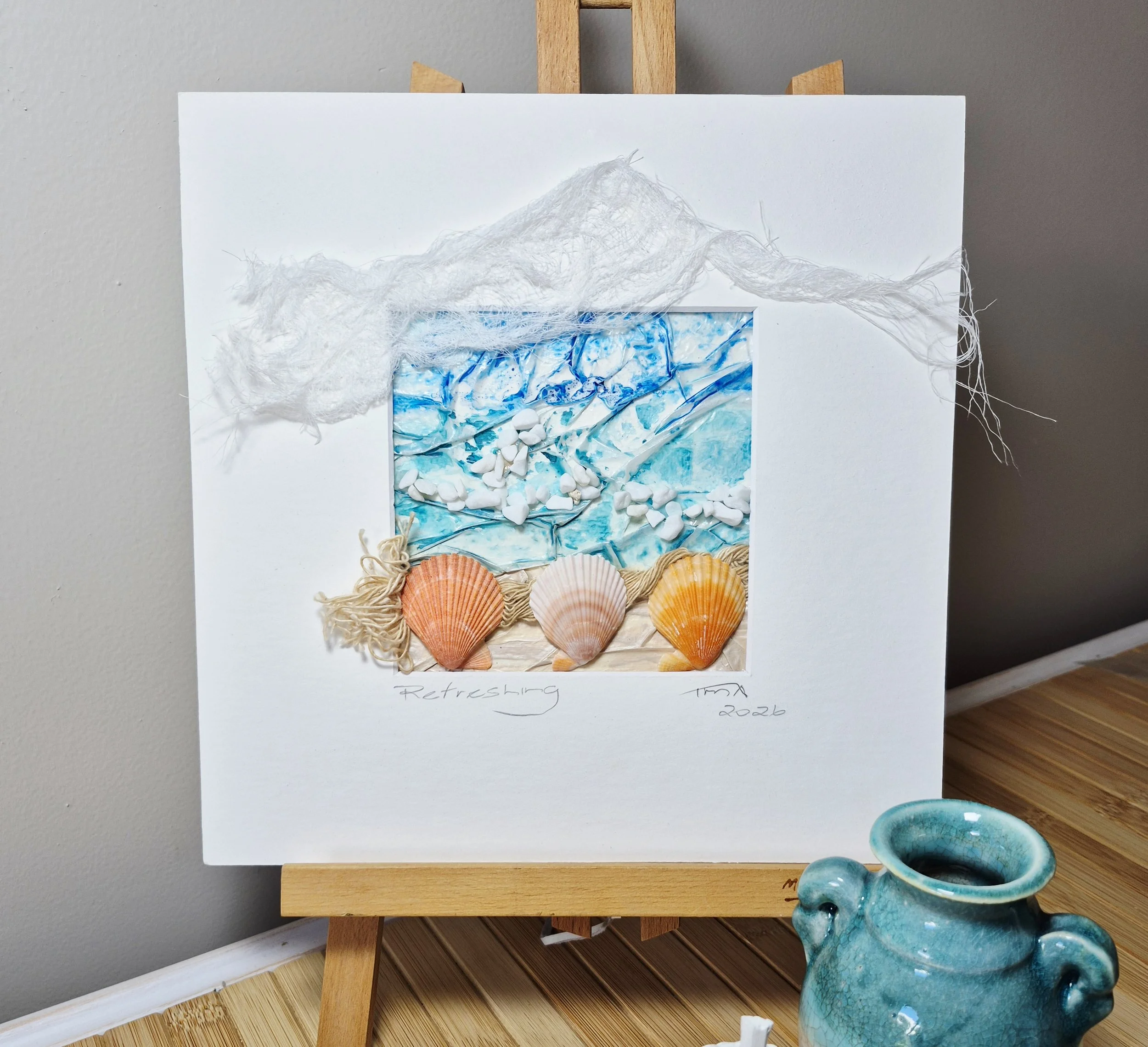 Miniature ocean scene with a blue marbled background, white stones, five orange shells, beige string, and a cotton-like cloud on white paper, handwritten notes and date.