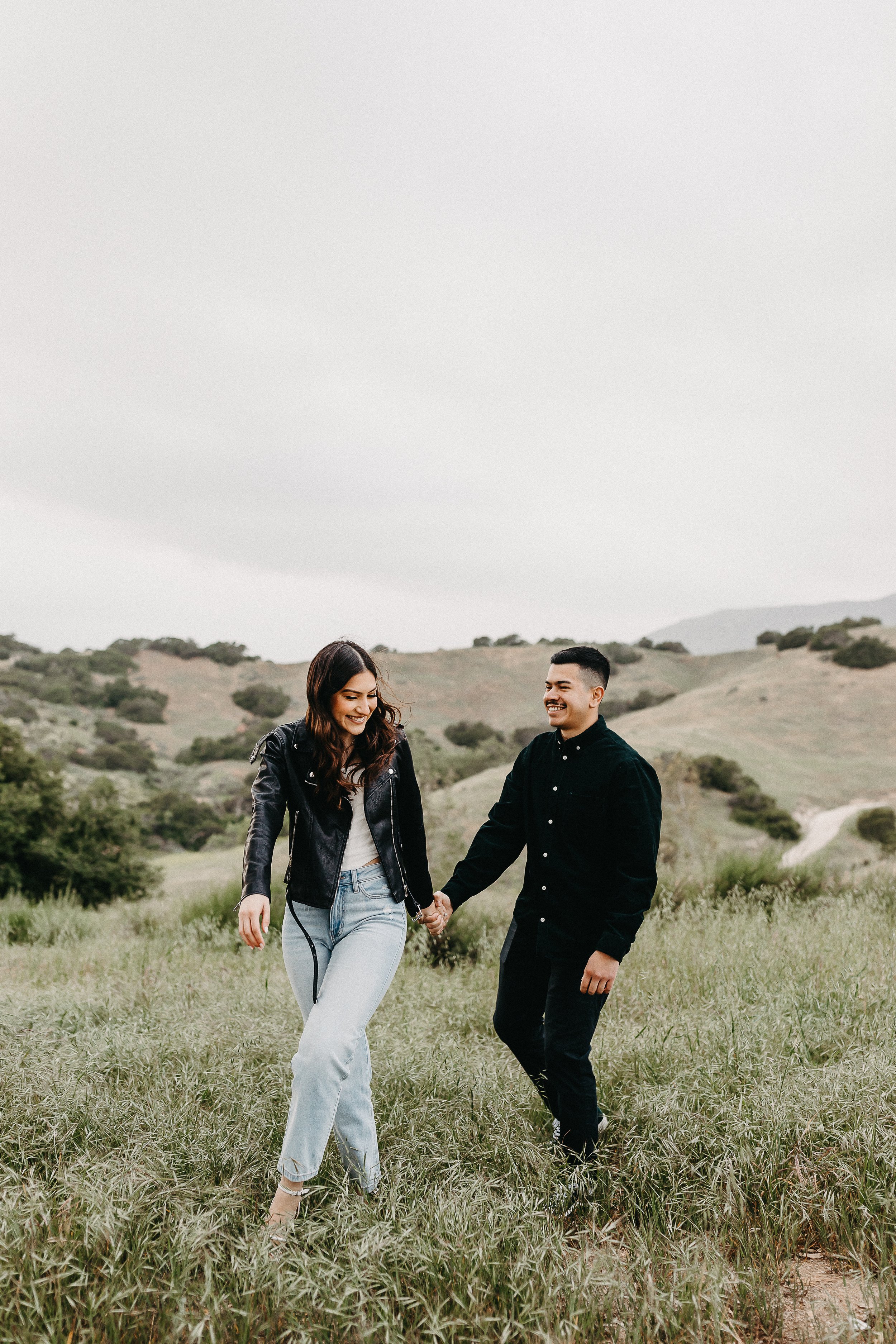 a women in a leather jacket and blue jeans with her fiancé in all black in a open green grass field for their engagement session
