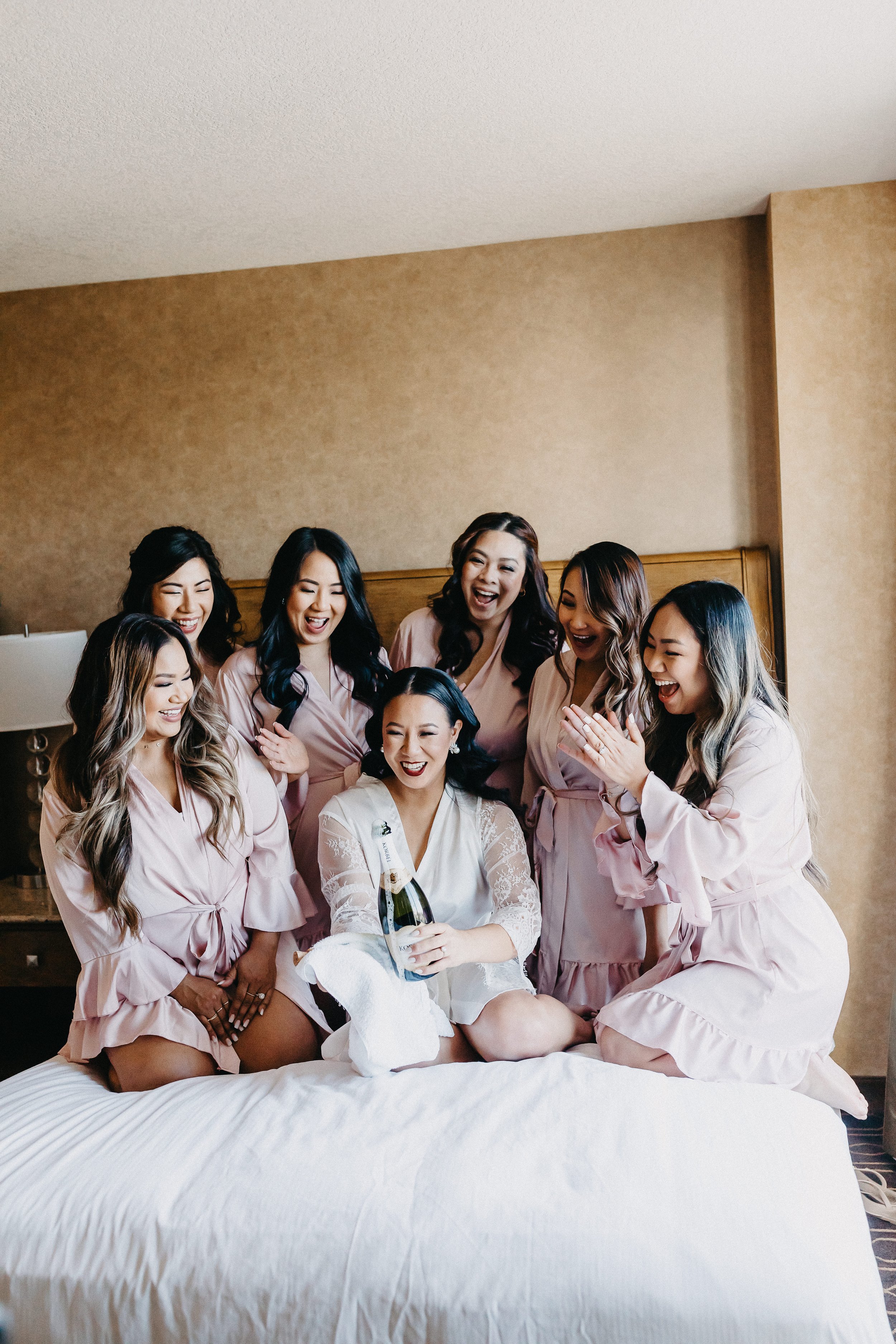 Bridesmaids in their robes on the bed cheering while the bride pops a champagne bottle.